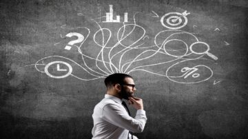 رجل يرتدي نظارات وقميص أبيض يقف بجانب جدار رمادي، يتأمل بجدية بينما تظهر فوق رأسه رسومات بيانية وأهداف وأسئلة، تعبر عن التفكير والتحليل.