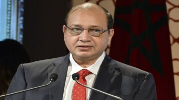 رجل يرتدي بدلة رسمية وقميص أبيض مع ربطة عنق حمراء، يتحدث من منصة، مع خلفية تحمل علم المغرب. يظهر في الصورة تعبير جاد أثناء إلقاء خطاب أو تقديم عرض.