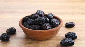 وعاء خشبي يحتوي على تمر أسود لامع، مع بعض حبات التمر متناثرة حول الوعاء، الخلفية خشبية.