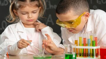 أطفال يجربون في المختبر، أحدهم يستخدم قطارة لإضافة سائل إلى طبق زجاجي، والآخر يرتدي نظارات واقية ويلاحظ التجربة. هناك أنابيب اختبار ملونة في الخلفية.
