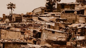 منظر لحي فقير يظهر منازل غير منظمة مبنية من مواد معاد تدويرها، تتكدس فوق بعضها البعض. العديد من أسطح المنازل مزودة بأطباق فضائية، وتحيط بها أشجار. تعكس الصورة ظروف المعيشة الصعبة في المناطق العشوائية.