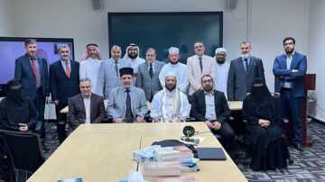 مجموعة من الرجال والنساء يرتدون ملابس تقليدية، يجلسون ويقفون حول طاولة في غرفة اجتماعات. يظهر في الصورة علماء دين وممثلون من خلفيات ثقافية متنوعة، مع وجود شاشات عرض في الخلفية. يعبّر المشهد عن التعاون والتفاعل في بيئة مهنية.