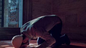 رجل مسلم يؤدي الصلاة في مسجد، مع التركيز على الركوع. الإضاءة خافتة، مما يبرز تفاصيل السجادة والمكان.