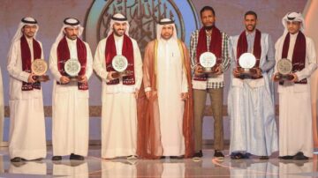 أشخاص يرتدون ملابس تقليدية يقفون على منصة احتفالية، يحملون جوائز. يظهر خلفهم شعار يتضمن نصوصاً عربية. المناسبة تتعلق بتكريم المتميزين في مجال معين.