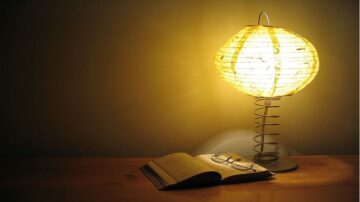 مصباح مضيء بتصميم دائري مصنوع من الورق، موضوع بجانب كتاب مفتوح ونظارات على طاولة خشبية. الإضاءة الدافئة تخلق جوًا مريحًا للقراءة.