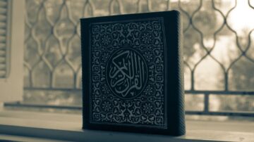 كتاب قرآن كريم مغلف بتصميم تقليدي، موضوع على حافة نافذة مع خلفية زجاجية مزخرفة. الضوء يتسلل من خلال الزجاج، مما يخلق جوًا هادئًا ومهيبًا.