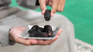 تمر طازج في طبق فضي، شخص يحمل حبة تمر واحدة بيده بينما تحتوي اليد الأخرى على طبق مليء بالتمور. الخلفية خضراء، مما يبرز جمال التمر.