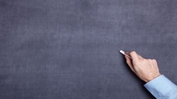 يد تحمل طبشورة بيضاء تكتب على سطح رمادي داكن.