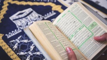 كتاب مفتوح يوضح صفحات تحتوي على نصوص باللغة العربية، مع تمييز أجزاء معينة باللون الأخضر. تظهر سجادة صلاة تقليدية في الخلفية، مما يعكس أجواء العبادة والدراسة الإسلامية.