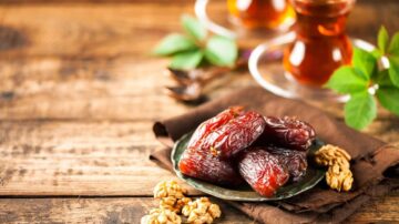 تمر طازج في صحن معدني مع جوز وشاي في أكواب زجاجية، موضوع على طاولة خشبية.