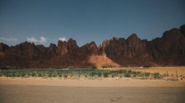 منظر طبيعي لجبال شديدة الانحدار بلون أحمر مائل للبني، يحيط بها أرض زراعية خضراء. السماء زرقاء مع بعض السحب البيضاء، والطريق الأسفلتي يمتد أمام الجبال. يعتبر هذا المشهد مثالاً رائعًا للمناظر الطبيعية الصحراوية.