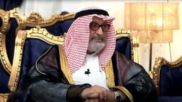 رجل مسن يرتدي ثوباً عربياً تقليدياً مع عقال وشماغ، يجلس على أريكة فاخرة ذات تصميم ذهبي في غرفة مضاءة بشكل دافئ.