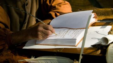 يد تظهر كتابة على ورقة مفتوحة باستخدام قلم ريش، مع وجود كتاب مغلق وأوراق بيضاء حوله. المشهد يعكس أجواء الكتابة التقليدية.