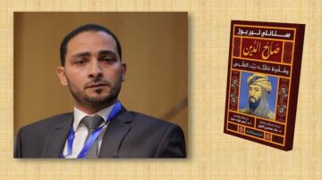 صورة لرجل يرتدي بدلة رسمية، يظهر بملامح جادة، بجانب كتاب يحمل عنوان "صلاح الدين" ويعرض صورة تاريخية لشخصية معروفة. الخلفية بسيطة مع لون فاتح، مما يبرز الكتاب والشخصية.