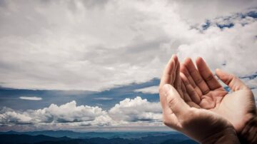 يدين مفتوحتين نحو السماء مع سحب بيضاء وكثيفة تتناثر في الخلفية الجبلية، تعكس مشاعر الأمل والتطلعات.