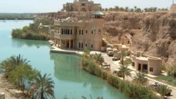 منظر خلاب لمبنى فاخر على ضفاف نهر، محاط بأشجار النخيل والتضاريس الصخرية. يظهر في الصورة أيضًا المركبات على طول الممر المائي.