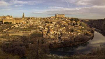 منظر بانورامي لمدينة طليطلة في إسبانيا، يظهر المعالم التاريخية والمباني القديمة على التلال، مع نهر يجري في الأسفل، وأجواء غائمة تضيف لمسة درامية للمنظر.