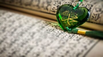 قلب أخضر شفاف يحمل اسم محمد مكتوب بالخط العربي، موضوع على صفحات من القرآن الكريم. يُظهر المشهد جمال الفن الإسلامي والروحانية.