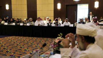 اجتماع رسمي في قاعة مؤتمرات، حيث يجلس عدد من الأشخاص حول طاولة مستديرة. يشارك الرجال والنساء في مناقشات، مع وجود زهور وزينة على الطاولة. يظهر أحد الحضور واقفًا في الخلفية.