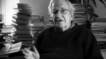 رجل مسن يرتدي سترة داكنة يجلس في غرفة مليئة بالكتب. تظهر خلفه أكوام من الكتب على الطاولة والجدران. يبدو في حالة تفكير وهو يتحدث، مع وجود إضاءة خفيفة تسلط الضوء على ملامح وجهه.