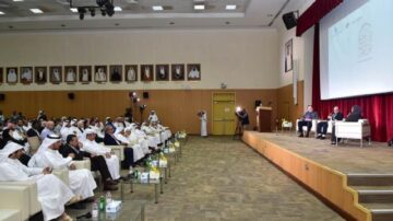 مؤتمر يجمع مجموعة من الحضور في قاعة كبيرة، حيث يجلس المتحدثون على المنصة أمام جمهور يتكون من رجال ونساء يرتدون الأزياء التقليدية. الجدار الخلفي مزين بصور شخصيات بارزة، والإضاءة تركز على المتحدثين.