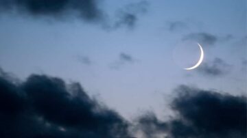 هلال رقيق يظهر في سماء زرقاء مع غيوم داكنة، مشهد ليلي هادئ يبرز جمال القمر في الفضاء.