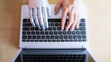 يد تظهر على لوحة مفاتيح كمبيوتر محمول بجوار يد روبوتية فضية، تعكس التفاعل بين الإنسان والذكاء الاصطناعي في البيئة الرقمية.