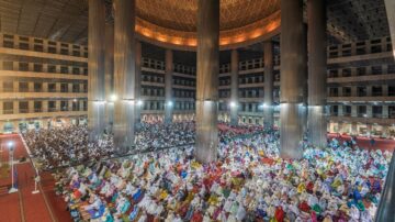 احتفال ديني حاشد في قاعة كبيرة، حيث يجلس الآلاف من الأشخاص في صفوف مرتبة. يرتدي المشاركون ملابس تقليدية متنوعة، ويتواجدون تحت أعمدة ضخمة وسقف مزخرف. الأضواء الساطعة تضيء المكان، مما يخلق أجواءً من الروحانية والتواصل الجماعي.