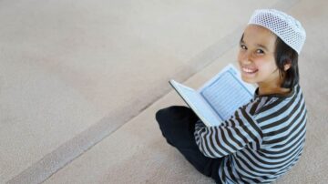 طفل صغير يجلس على الأرض يقرأ القرآن، يرتدي قبعة بيضاء وقميص مخطط باللونين الأسود والأبيض، مبتسم ويظهر سعادة أثناء الدراسة.