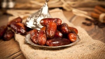 تمر طازج يوضع في طبق فضي على قماش خشن. تتميز التمور بلونها البني الداكن وقوامها اللزج، مع خلفية من الخشب لإضافة لمسة طبيعية.