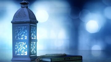 فانوس مزخرف مضاء بجانب مصحف وسبحة، مع خلفية زرقاء ضبابية. يعكس المشهد أجواء روحانية تعبر عن التقاليد الإسلامية.