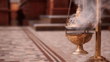 مبخرة ذهبية تدخن في مكان داخلي، مع تفاصيل معمارية خشبية وأرضية مزخرفة. تبرز البخور المتصاعد من المبخرة الأجواء الروحية والاحتفالية.