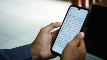 رجل يحمل هاتفًا ذكيًا يعرض نصًا باللغة العربية. النص يبدو كأنه جزء من كتاب أو وثيقة. الخلفية غير واضحة، مما يركز الانتباه على الشاشة.