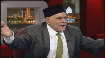 رجل مسن يرتدي بدلة رسمية وقبعة، يتحدث بحماسة أثناء ظهور تلفزيوني. الخلفية تحتوي على إضاءة ليلية لمدينة.