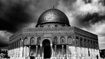 قبة الصخرة في القدس، مع قبتها الذهبية الأيقونية، تحت سماء غائمة. يظهر البناء بتفاصيله المعمارية الفريدة، بينما يتواجد زوار حوله.