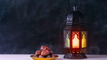فوانيس رمضان مزخرفة تصدر ضوءًا دافئًا بجانب طبق يحتوي على التمر. الخلفية داكنة، مما يبرز جمال الفانوس والتمر.