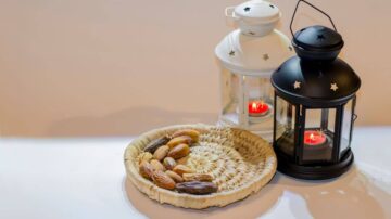 سلة مصنوعة من القش تحتوي على أنواع مختلفة من التمر، بجانب فوانيس بيضاء وسوداء تحمل نجومًا، مع شمع مضاء داخل كل فانوس.