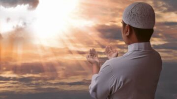 رجل يرتدي قميصًا أبيض وقبعة تقليدية، يقف متجهًا نحو السماء، يرفع يديه للدعاء تحت أشعة الشمس المتسللة عبر الغيوم. المنظر يعكس لحظة من الروحانية والسكينة.