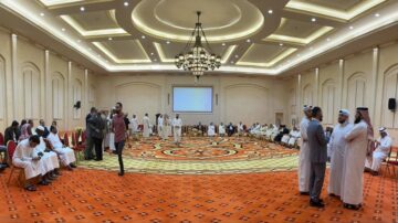 اجتماع كبير في قاعة مزينة بألوان دافئة، حيث يجلس الحضور على كراسي مرتبة بشكل دائري. يرتدي المشاركون ملابس تقليدية، ويبدو أنهم يتبادلون الحديث. تركز الإضاءة على الشاشة الكبيرة في مقدمة القاعة، مما يضيف جوًا رسميًا للمناسبة.