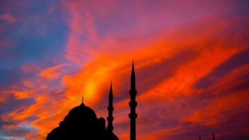 غروب شمس ملون خلف مسجد مع مآذن بارزة في السماء، تظهر تفاصيل السحب بألوان دافئة من البرتقالي والأرجواني.