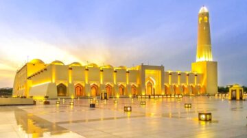 مسجد معمارية رائعة مضاءة بأضواء دافئة، مع مئذنة مميزة، في سماء مغربية خلال غروب الشمس. الأرض مبللة وتعكس الأضواء، مما يخلق جوًا ساحرًا.