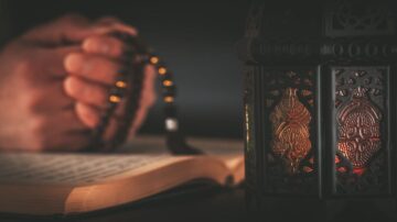 يد تضم حبات مسبحة بجوار مصباح مزخرف، مع كتاب مفتوح في الخلفية. تعكس الأضواء الدافئة من المصباح أجواءً روحية وهادئة.