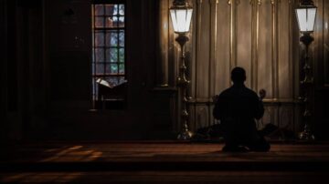 رجل يؤدى الصلاة في مسجد مظلم، مع إضاءة خافتة من مصابيح جانبية، يركز في لحظة تأمل روحي.