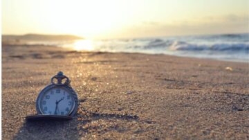 ساعة جيب قديمة موضوعة على رمال الشاطئ مع غروب الشمس في الخلفية، الأمواج تتلاطم بلطف على الساحل.