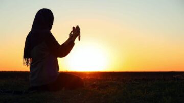 امرأة ترتدي حجابًا تجلس في العشب أثناء غروب الشمس، ممسكة بمسبحة في يديها. الخلفية تُظهر شمس الغروب في الأفق، مما يخلق مشهدًا هادئًا وملهمًا.