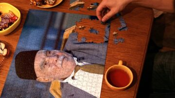 شخص يجمع قطع بانوراما على طاولة خشبية، حيث يظهر جزء من صورة لرجل يرتدي قبعة. بجانب البازلت، توجد أطباق صغيرة تحتوي على حلويات وكوب من الشاي.