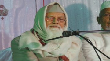 رجل مسن ذو لحية بيضاء يرتدي غطاء رأس تقليدي وشال، يجلس بجانب رجل آخر في حدث ثقافي، مع ميكروفون أمامه وخلفية مزينة بالقماش.