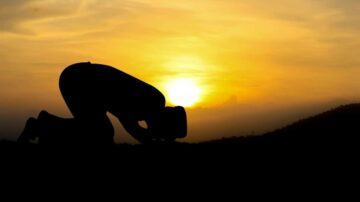 صورة ظلية لشخص يؤدي الصلاة في اتجاه الشمس الغاربة، مع سماء داكنة تتداخل فيها درجات البرتقالي والأصفر. تعبر الصورة عن السلام والروحانية في لحظة هادئة.