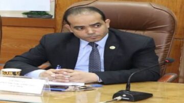 رجل أعمال يرتدي بدلة رسمية، يجلس على مكتب أثناء اجتماع. يظهر في الصورة تعبير جاد مع تركيز على المناقشة، مع ملاحظات وأدوات مكتبية حوله.