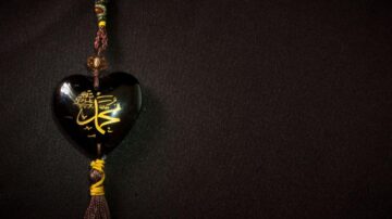 قلادة على شكل قلب بلون أسود، مزينة بنقوش ذهبية. تحتوي على خيوط ملونة وتفاصيل متقنة، موضوعة على خلفية داكنة.